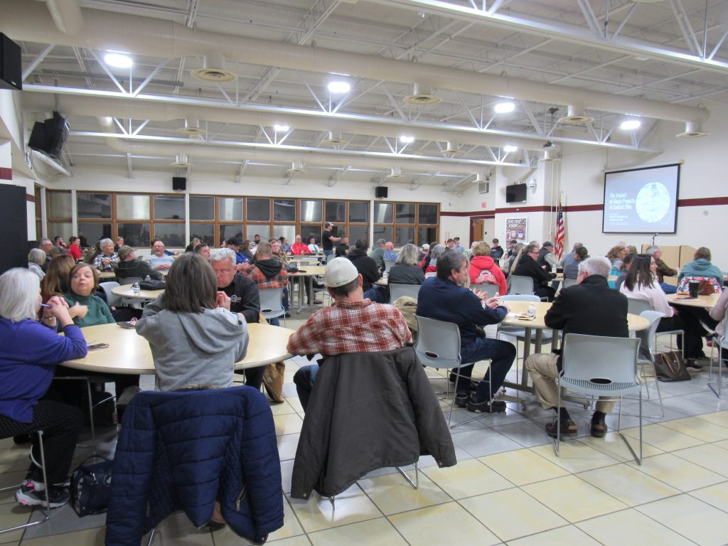 a room full of people sitting at cafeteria tables listening to Betsy Goldstein talk about Intel's mega project